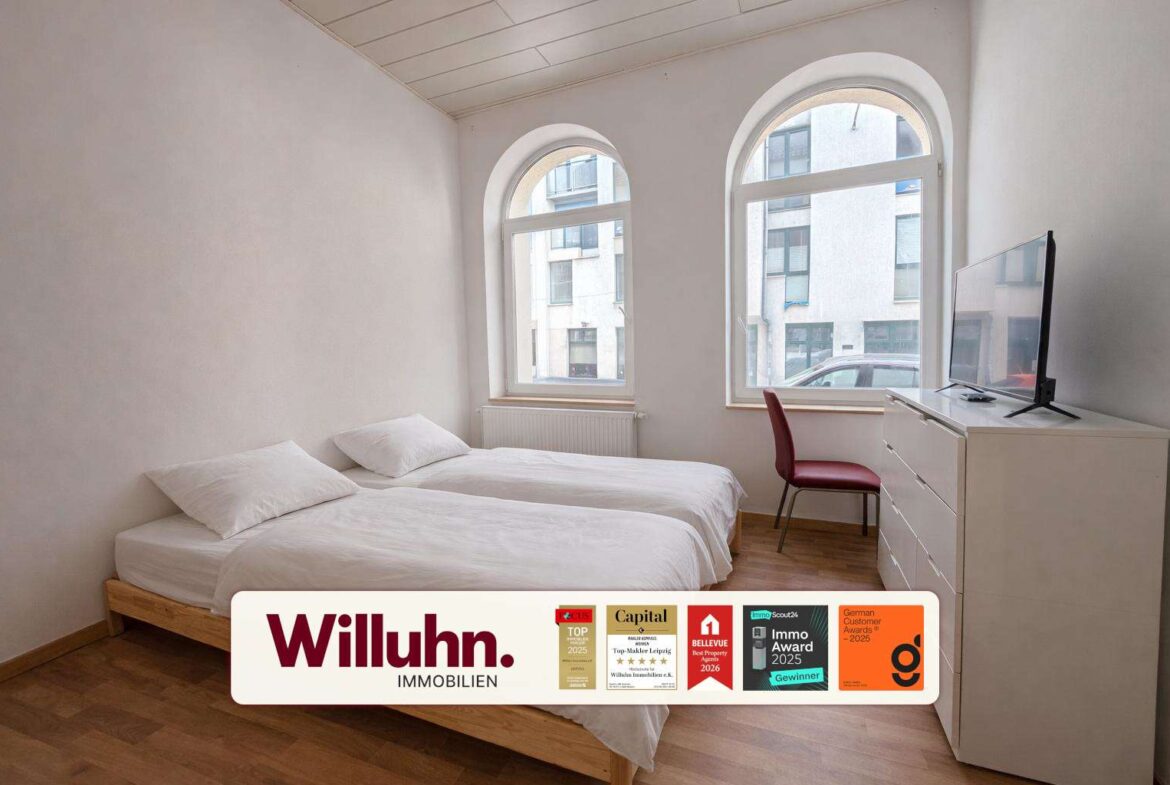 Bright apartment bedroom with two single beds, white bedding, a white dresser with a flat-screen TV, and a red chair by large arched windows.
