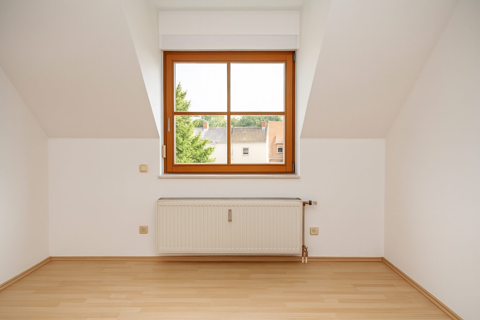 Bright attic room with a wooden-framed window, white walls, and a white radiator beneath; light hardwood floor.