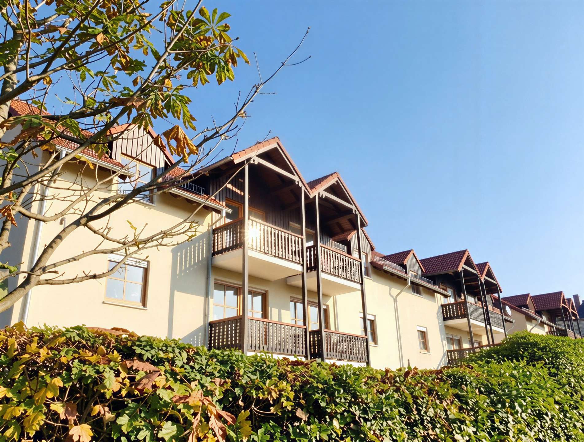 Residential building with multiple dark wood balconies and red-tiled roofs against a clear blue sky, with a hedge in the foreground and a tree partially framing the scene.