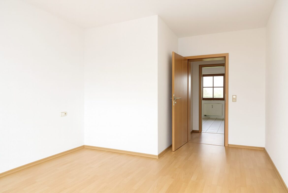 Empty room with light wood flooring and white walls; an open wooden door reveals a second room with a window and radiator.