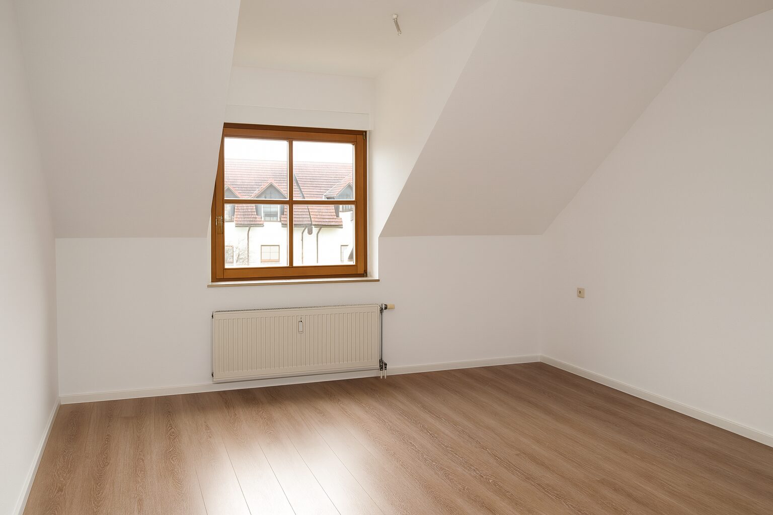 A bright empty attic room with white walls, a wooden-framed window, and a radiator under the window; light wood flooring shows through.