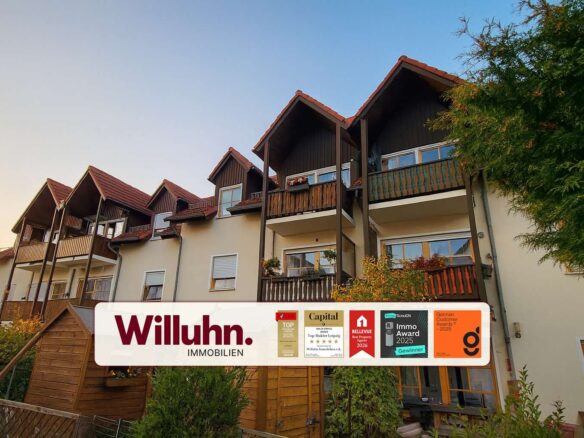 Apartment building with multiple brown wooden balconies and red-tiled roofs under a blue sky, overlaid by a Willuhn Immobilien banner.