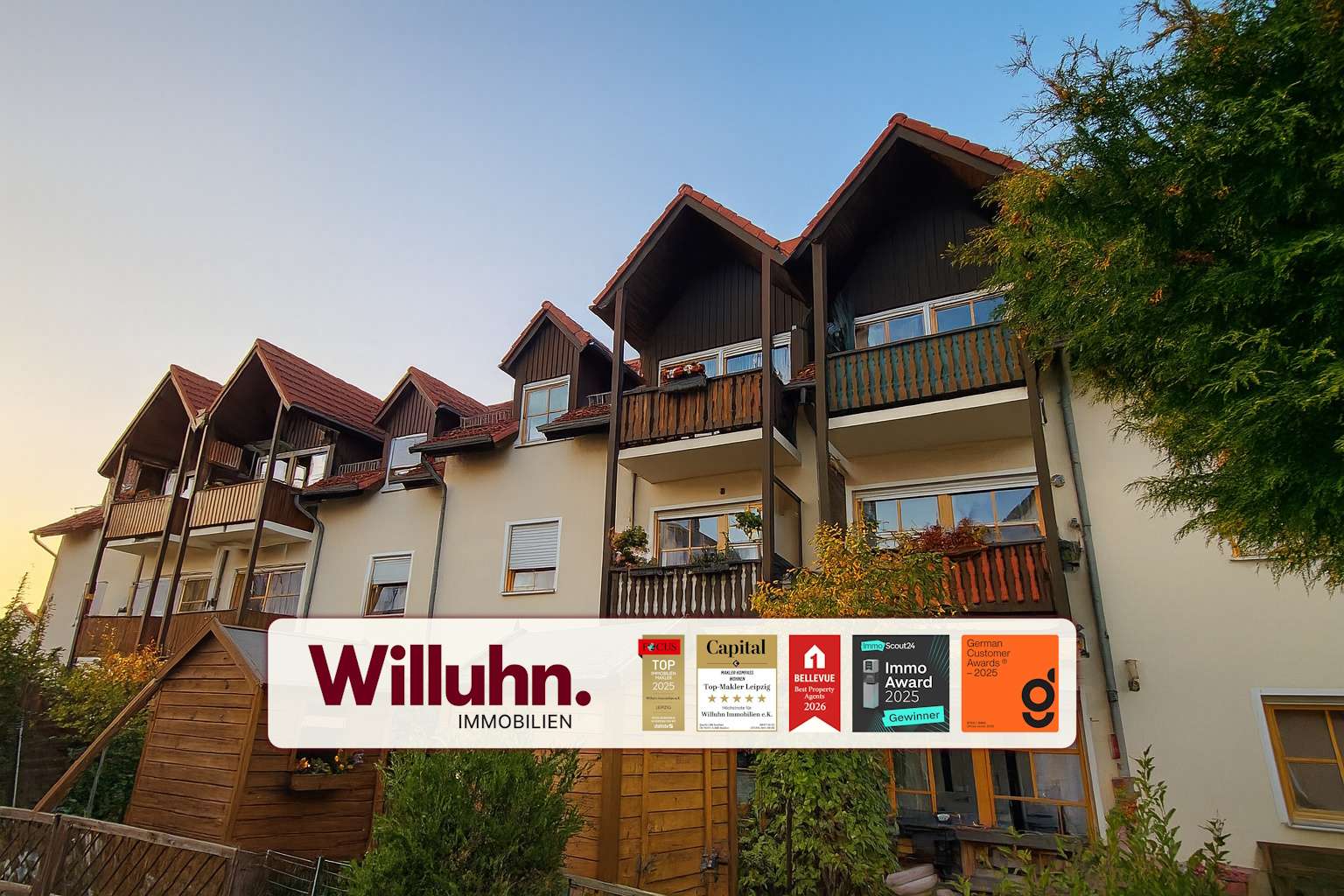 Apartment building with multiple brown wooden balconies and red-tiled roofs under a blue sky, overlaid by a Willuhn Immobilien banner.
