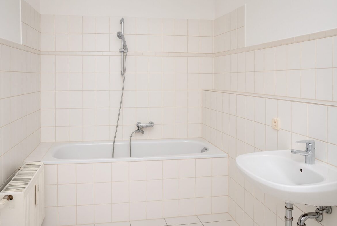 Bright white-tiled bathroom with a rectangular bathtub, handheld shower, and chrome fixtures nearby a sink and radiator