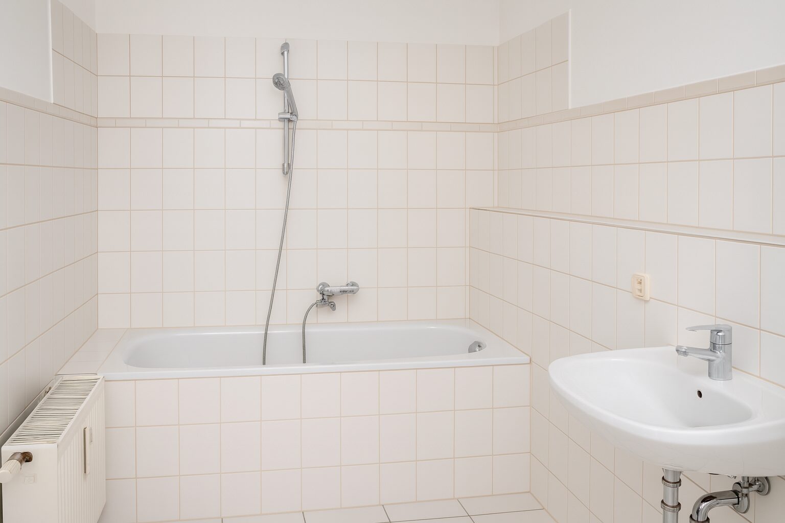 Bright white-tiled bathroom with a rectangular bathtub, handheld shower, and chrome fixtures nearby a sink and radiator