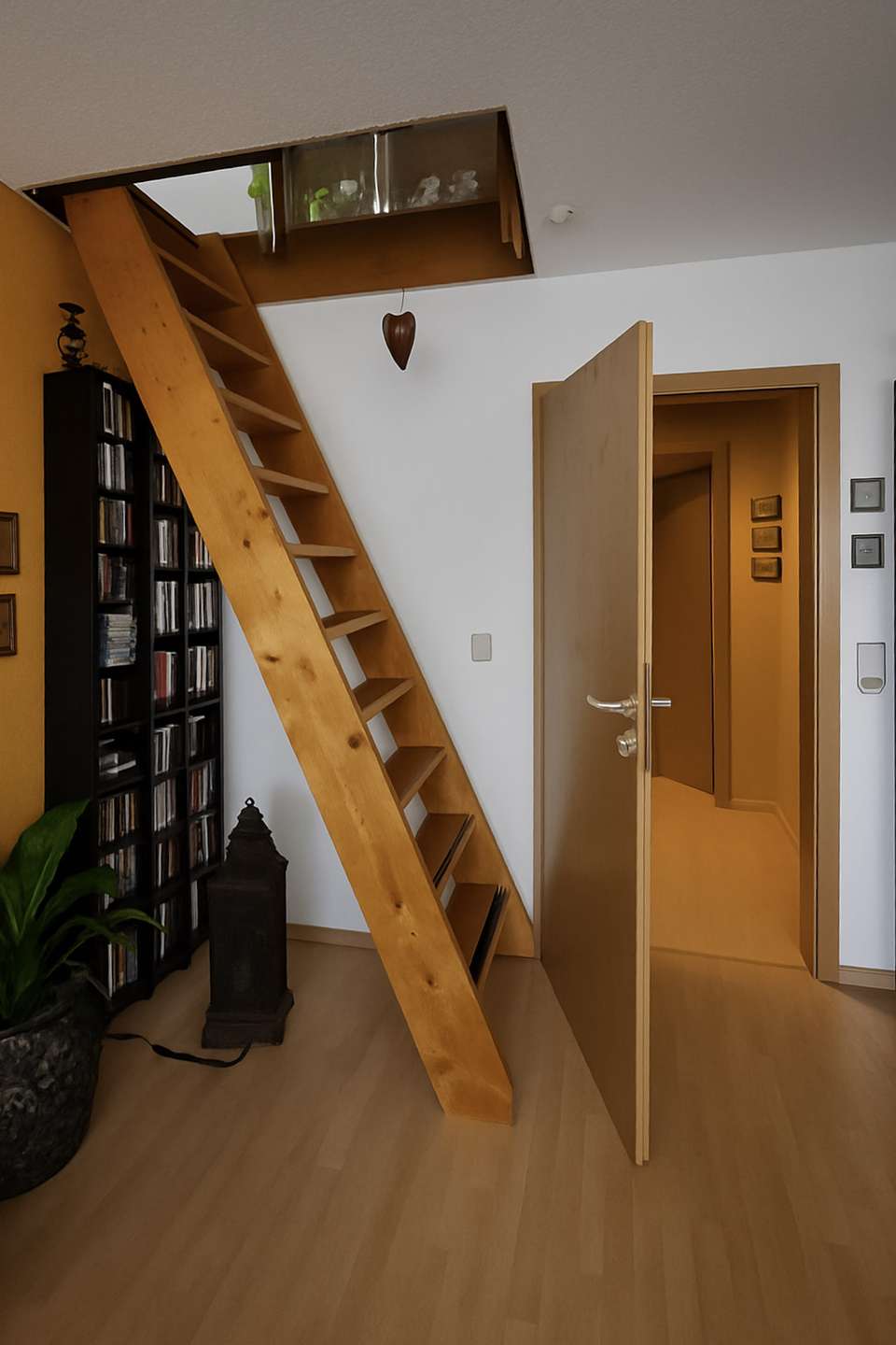 Wooden diagonal loft ladder leading to a small upper hatch opening in a cozy living area with bookshelves and a door ajar to another room