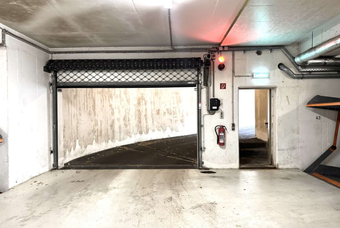 Industrial loading bay interior with large overhead rolling door and chain-link protection, concrete walls, and exposed pipes; a fire extinguisher and exit sign mounted on a column.