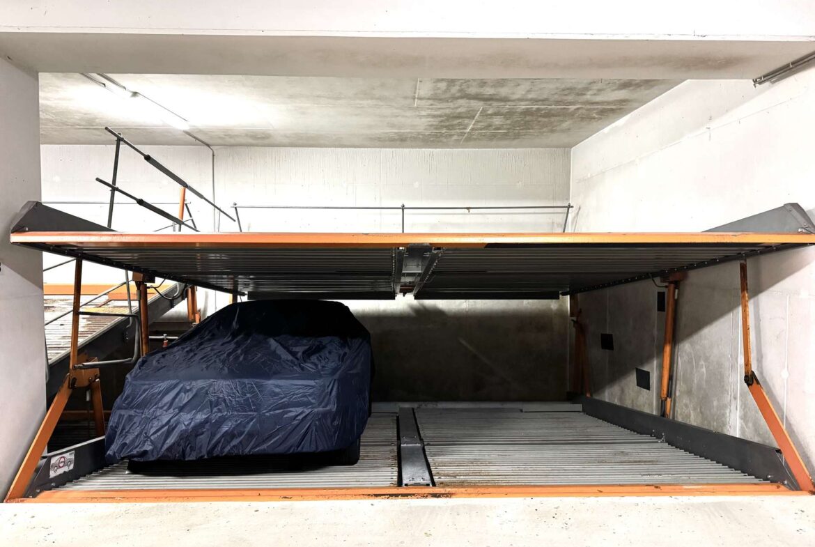 Blue tarp-covered car on an orange-framed car lift in a dim concrete garage setting, with metal rails above.