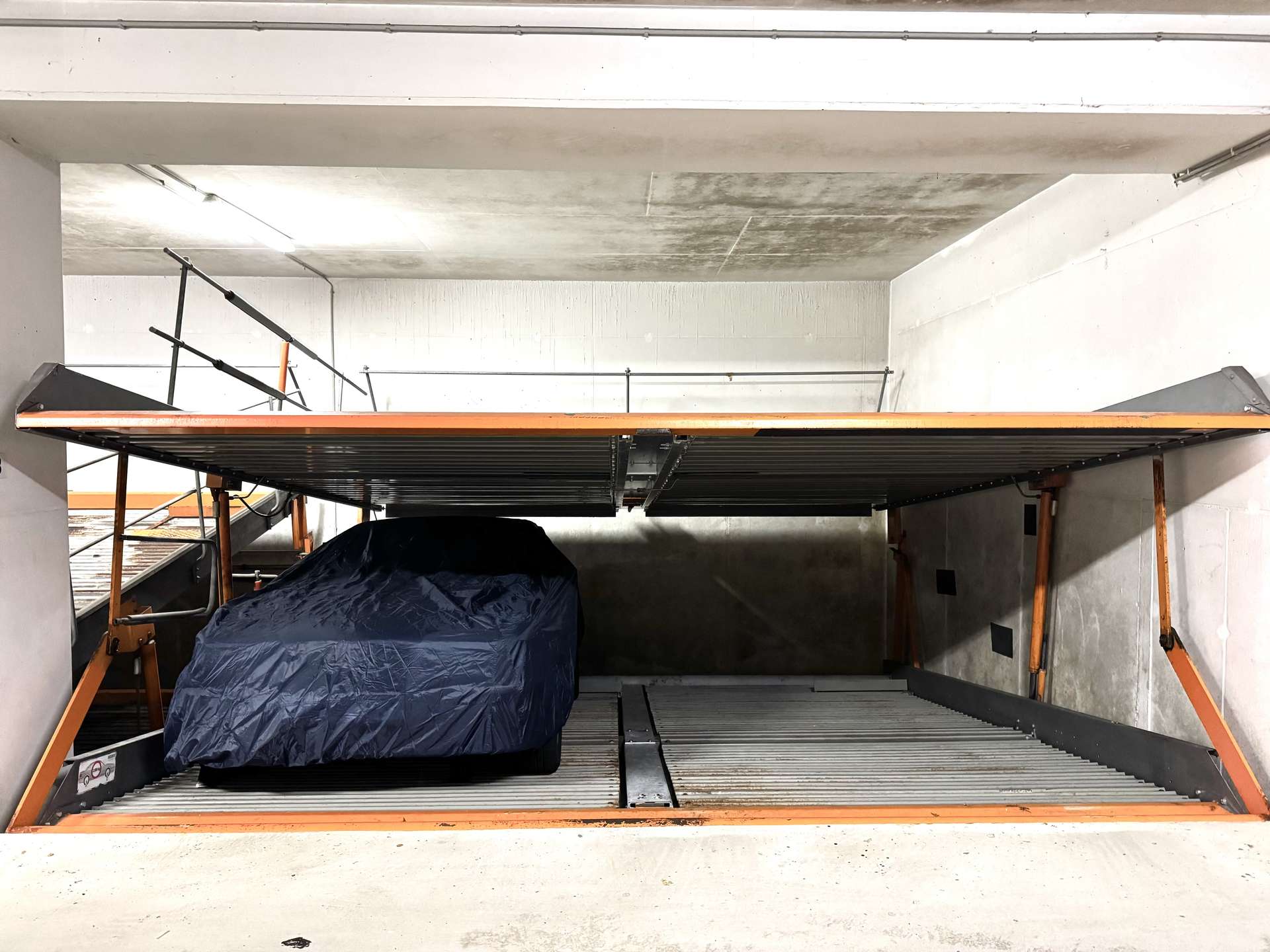 Blue tarp-covered car on an orange-framed car lift in a dim concrete garage setting, with metal rails above.