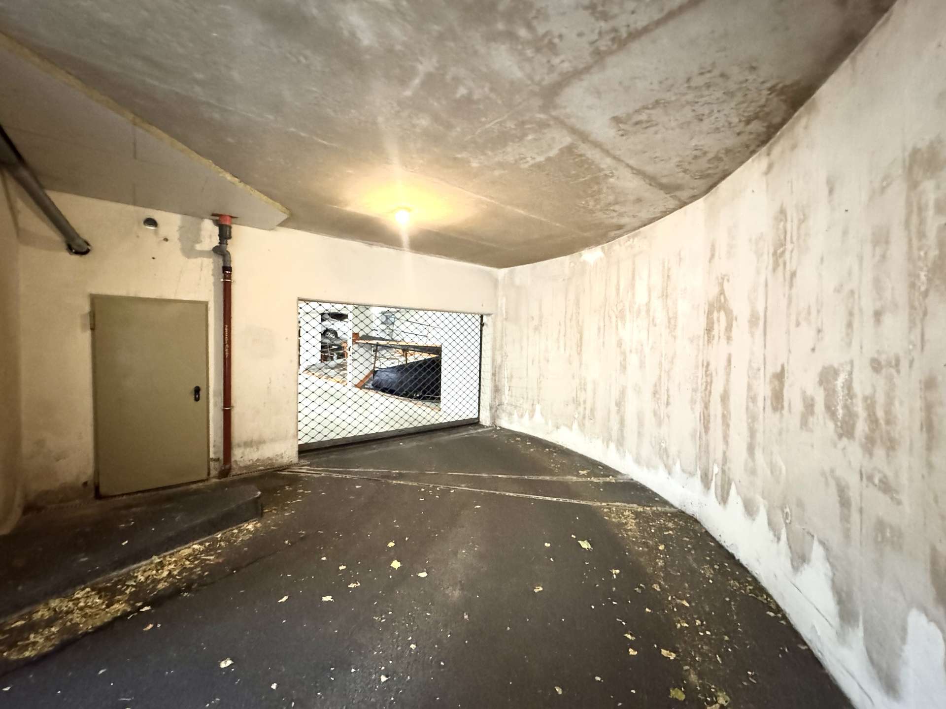 Industrial underground parking area with curved ceiling, bare white walls, a door on the left, exposed red pipe, and a mesh window showing another space beyond.