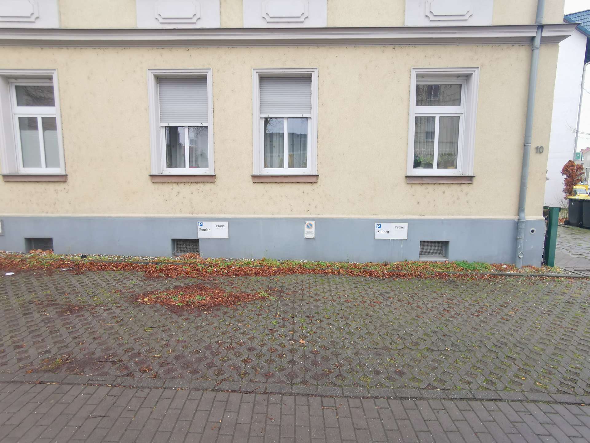 Beige building facade with three white-framed windows above a grey base, on a cobblestone sidewalk with fallen leaves in autumn colors.