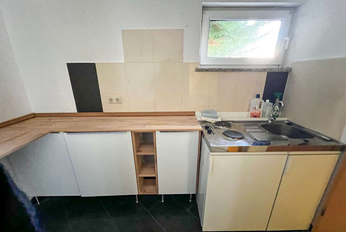 Compact kitchenette with light wood countertops, white cabinets, sink, and a two-burner electric stove under a window with beige tile backsplash.