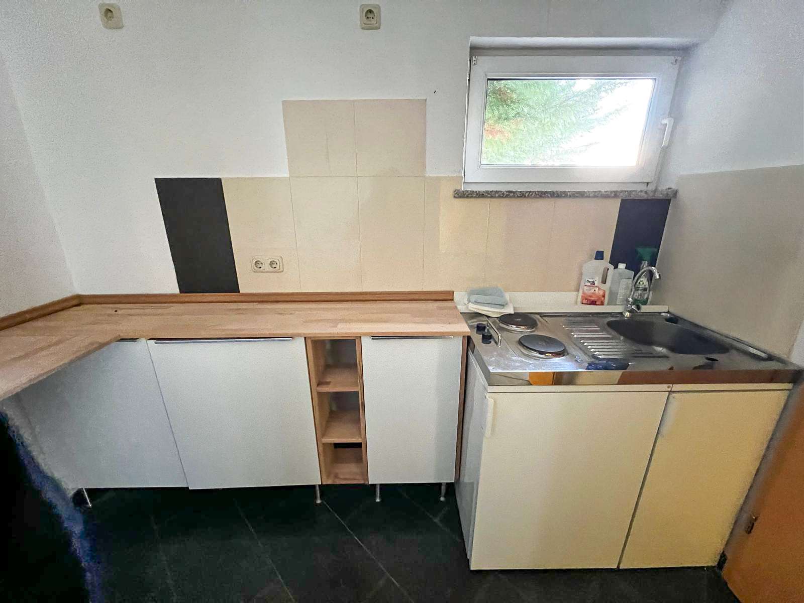 Compact kitchenette with light wood countertops, white cabinets, sink, and a two-burner electric stove under a window with beige tile backsplash.