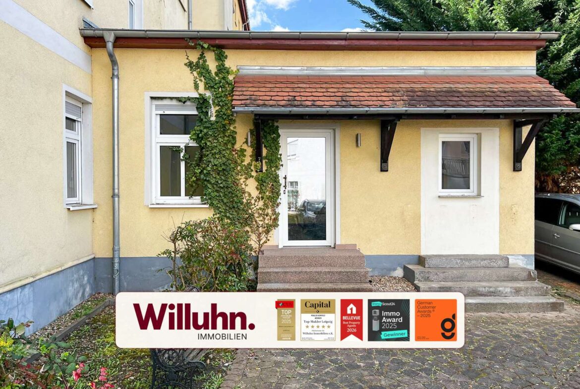 Front entrance of a yellow house with white trim, ivy climbing the wall, steps leading to a glass door, and a Willuhn Immobilien sign with awards in the foreground.