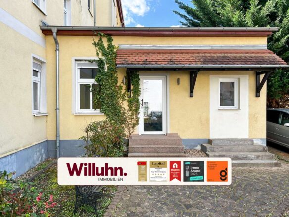 Front entrance of a yellow house with white trim, ivy climbing the wall, steps leading to a glass door, and a Willuhn Immobilien sign with awards in the foreground.