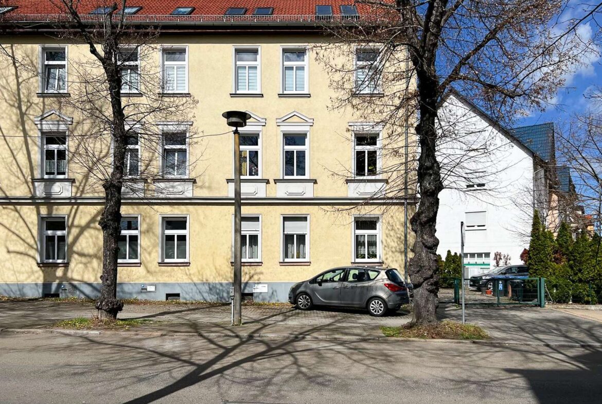 Yellow three-story residential building with white-framed windows, leafless trees in front, and a silver hatchback parked along the curb on a sunny day.