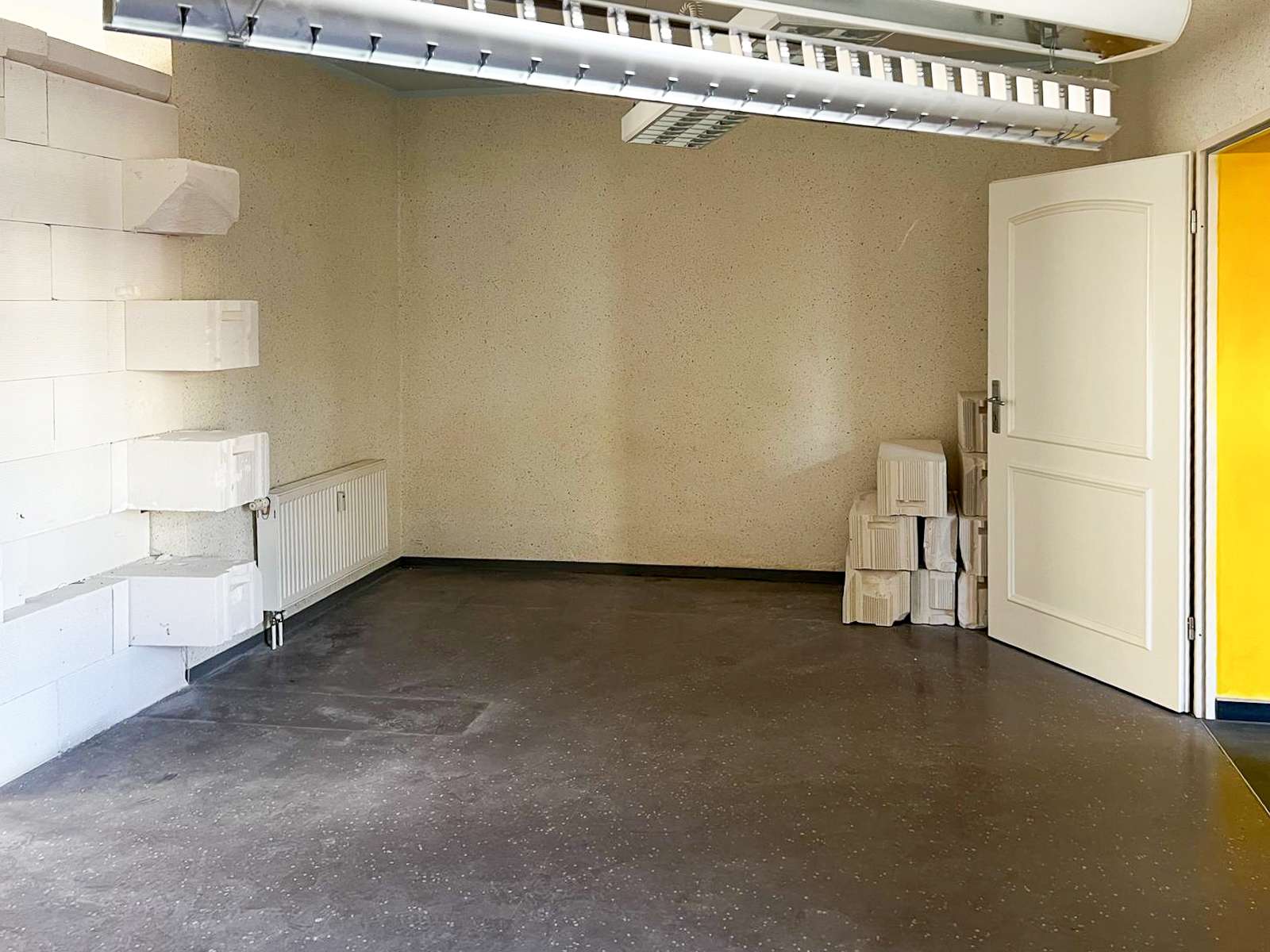 A small empty storage room with white stacked blocks along the left wall and a radiator beneath them, an open door on the right, and a pile of white blocks by the door.