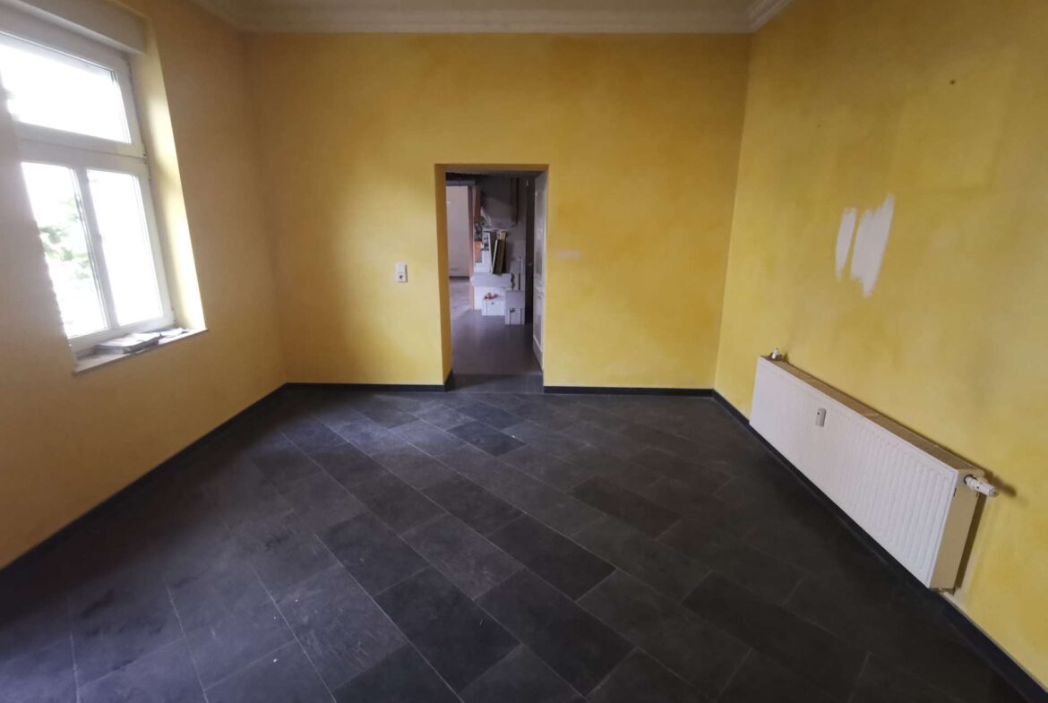 Yellow-painted empty room with dark tile floor, radiator on the right, window on the left, and a doorway leading to another room under renovation.