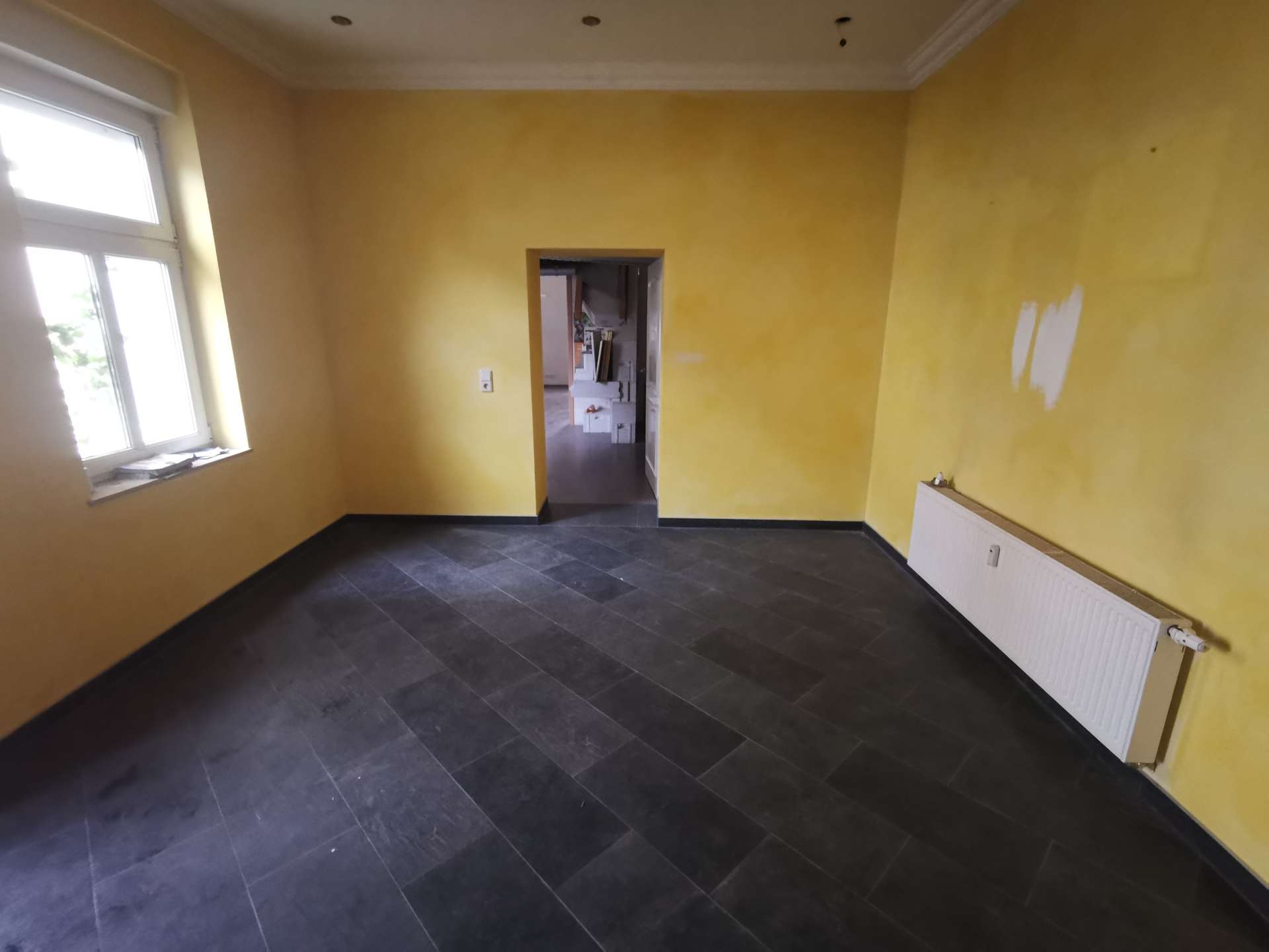 Yellow-painted empty room with dark tile floor, radiator on the right, window on the left, and a doorway leading to another room under renovation.