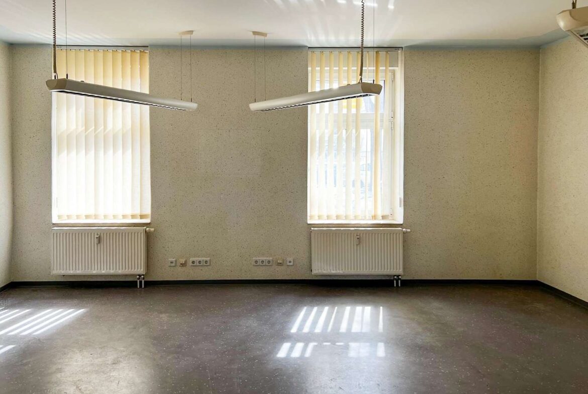 Empty, unused room with two tall windows and vertical blinds, radiators beneath each window, and suspended linear light fixtures.