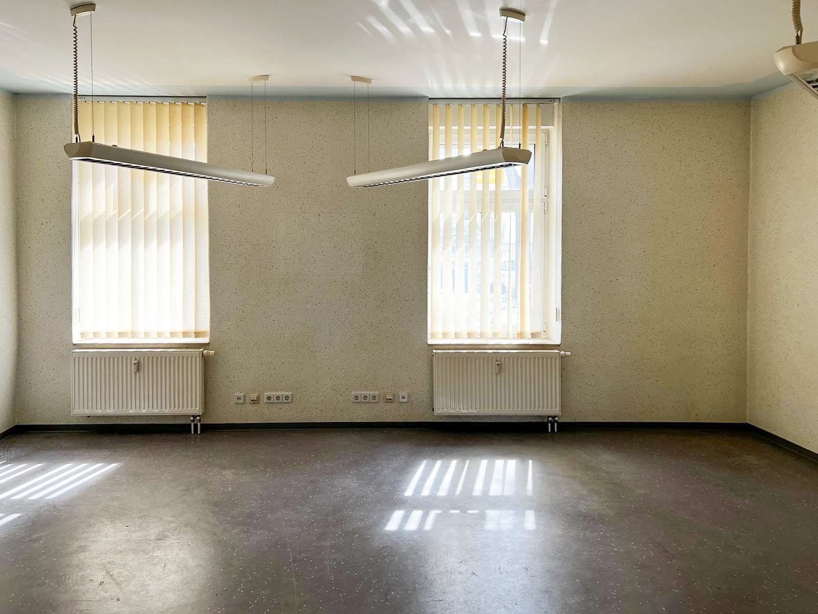 Empty, unused room with two tall windows and vertical blinds, radiators beneath each window, and suspended linear light fixtures.