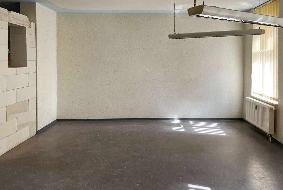 Empty office room with speckled beige walls, dark speckled floor, and a sunlit window with vertical blinds on the right; radiator beneath the window.