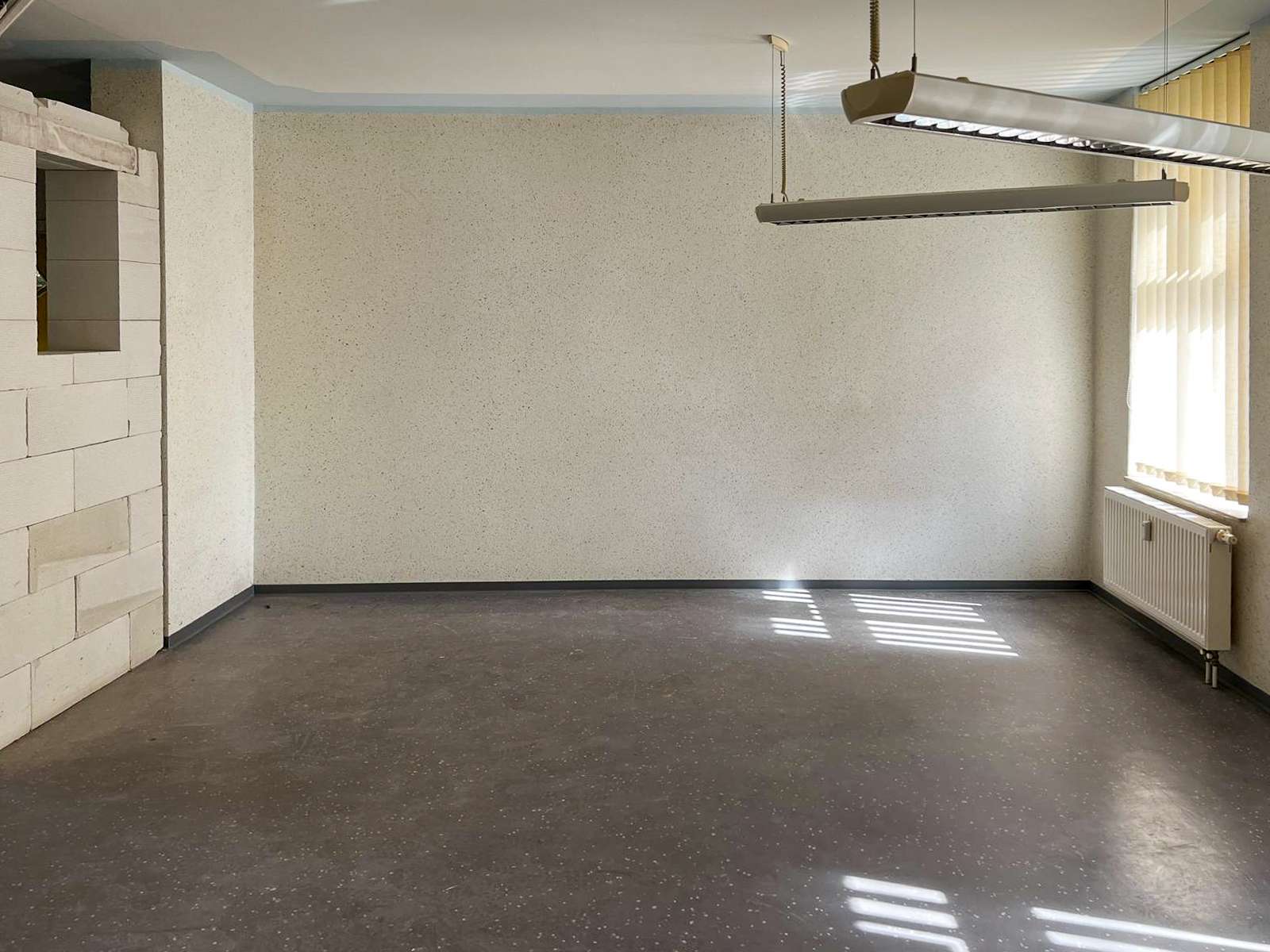 Empty office room with speckled beige walls, dark speckled floor, and a sunlit window with vertical blinds on the right; radiator beneath the window.