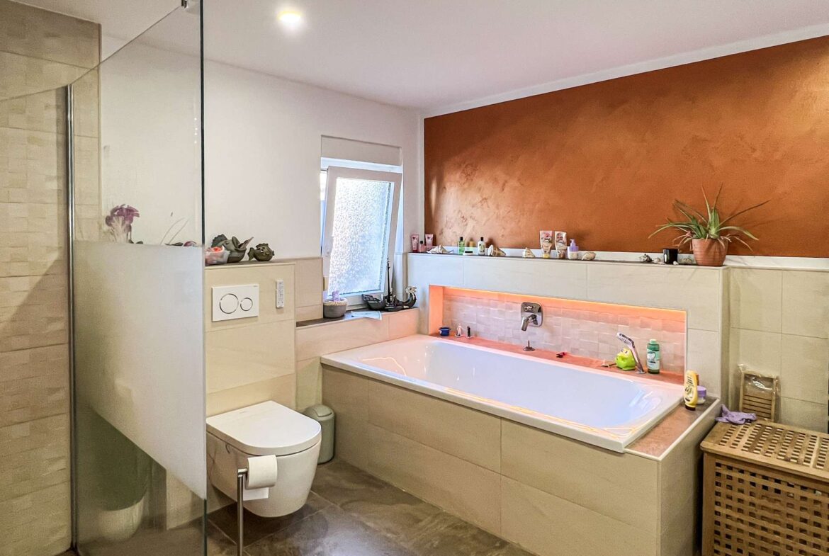 Modern bathroom with glass shower, built-in tub, orange accent wall, and a potted plant on the ledge; toiletries arranged along the shelf.