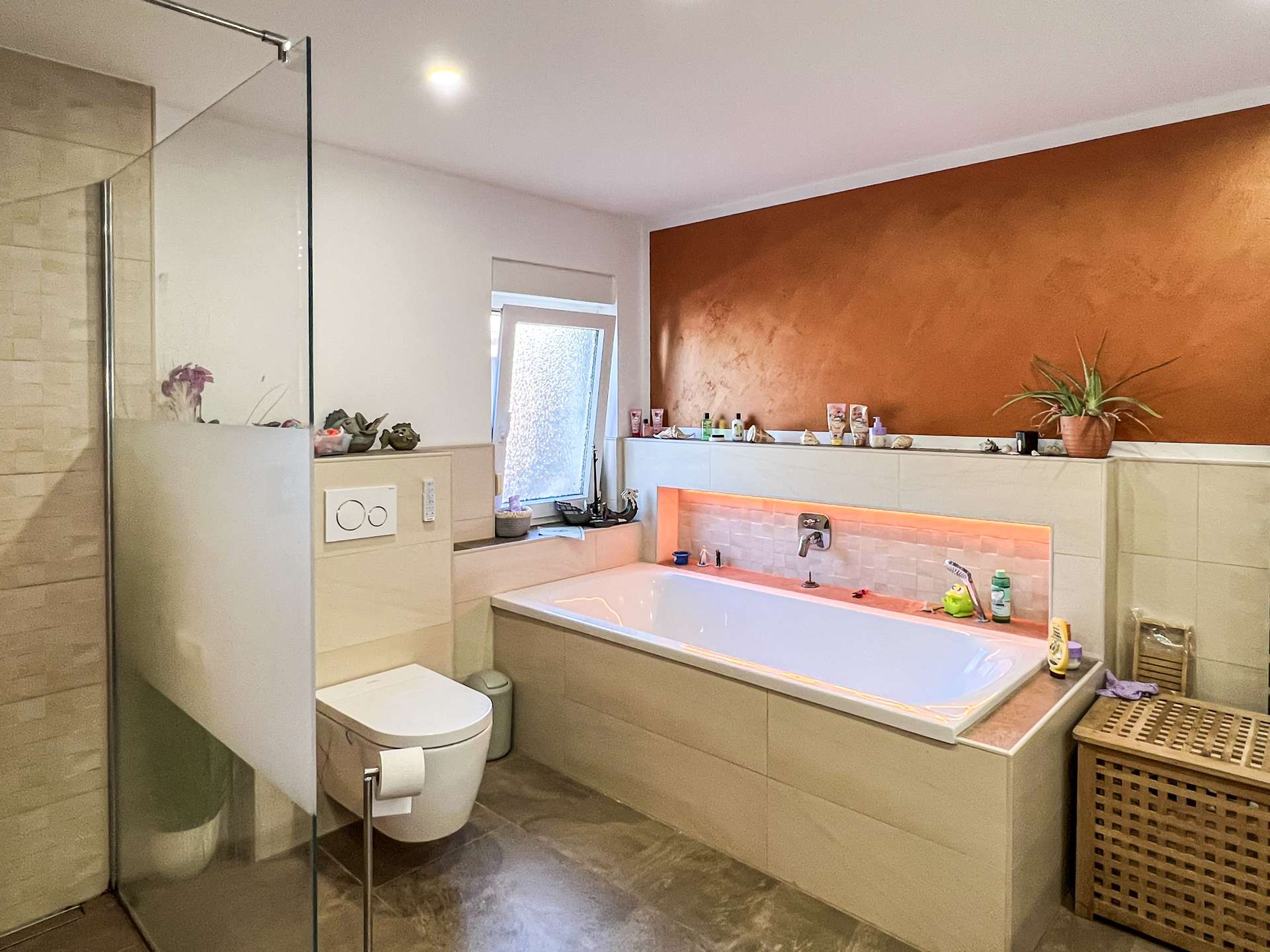 Modern bathroom with glass shower, built-in tub, orange accent wall, and a potted plant on the ledge; toiletries arranged along the shelf.
