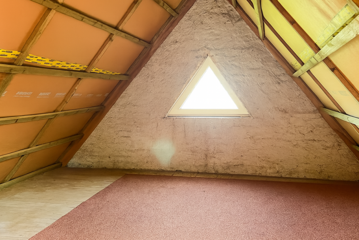 Attic with a triangular window in the gable wall, orange insulation, exposed wooden beams, and a pink carpeted floor.