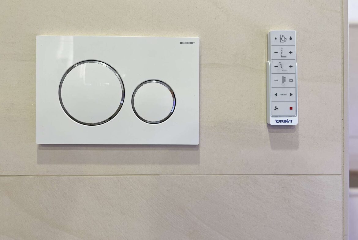 Wall-mounted Geberit flush plate with two large chrome circular buttons next to a vertical Duravit control panel on a tiled bathroom wall.