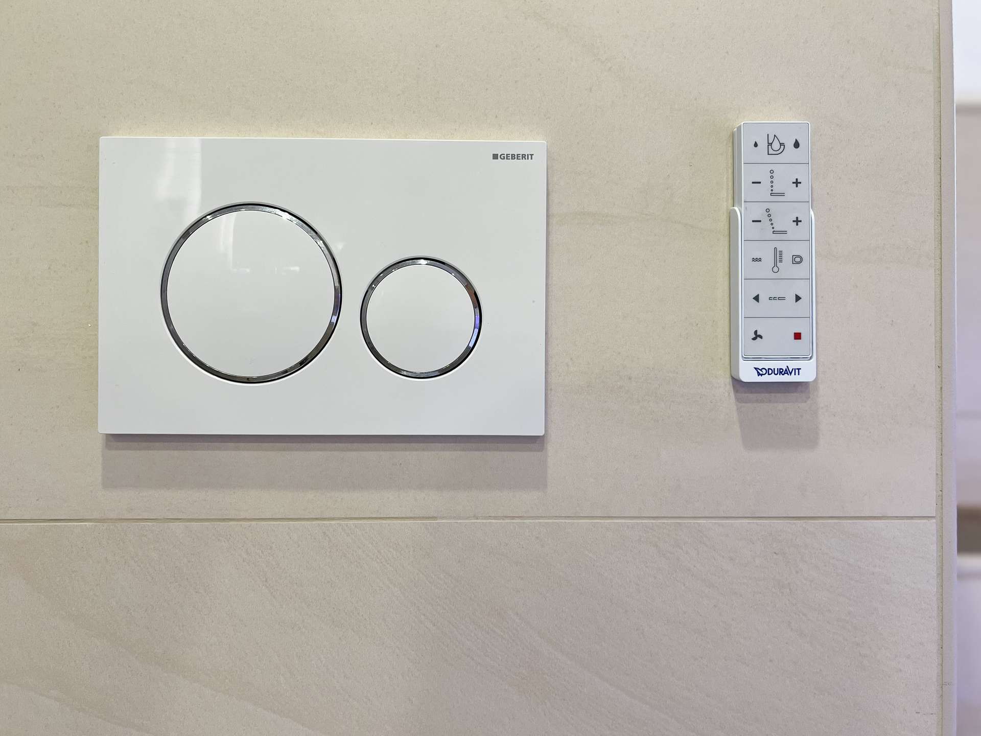 Wall-mounted Geberit flush plate with two large chrome circular buttons next to a vertical Duravit control panel on a tiled bathroom wall.