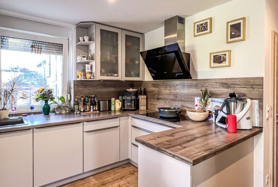 Modern kitchen with grey lower/upper cabinets, wooden countertops, and a black angled range hood over the stove. A window with plants and shelf items on the left adds light.