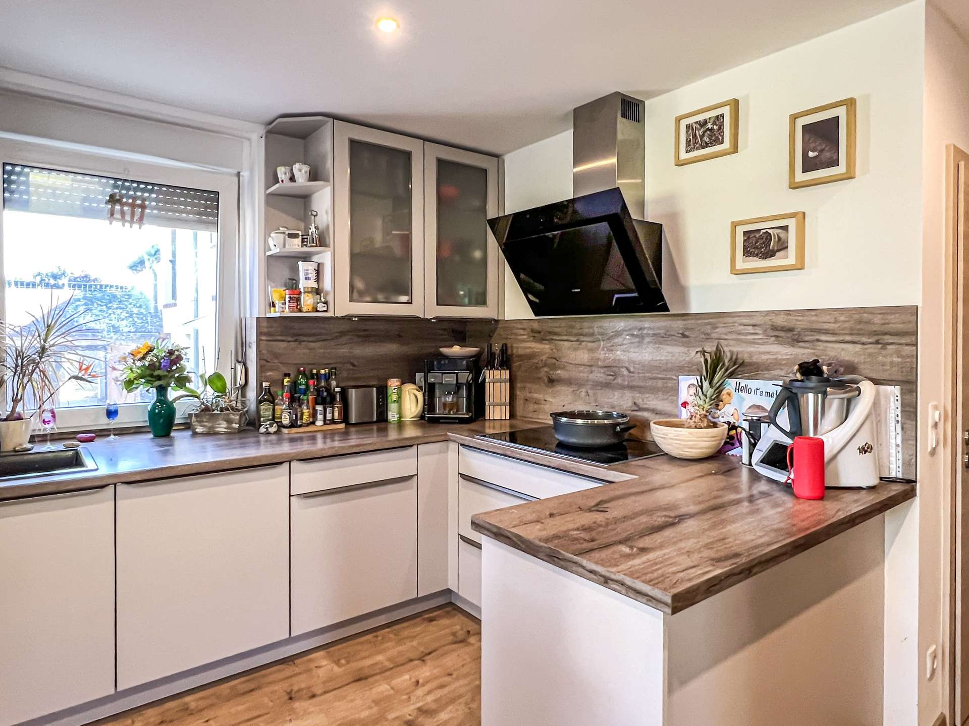 Modern kitchen with grey lower/upper cabinets, wooden countertops, and a black angled range hood over the stove. A window with plants and shelf items on the left adds light.