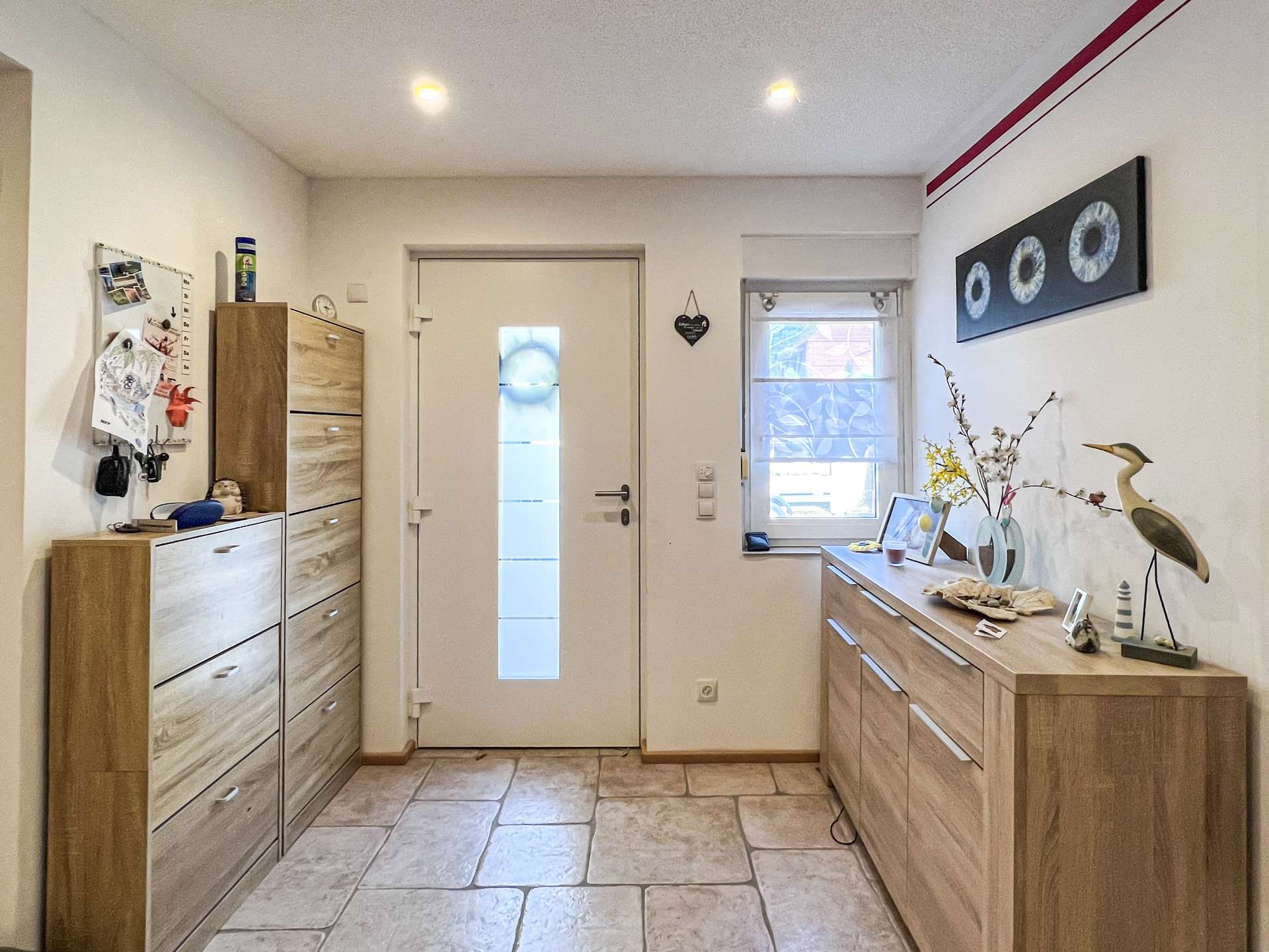 Bright entryway with light wood storage units lining both sides, a white door with a vertical frosted panel, and a dresser topped with decor.