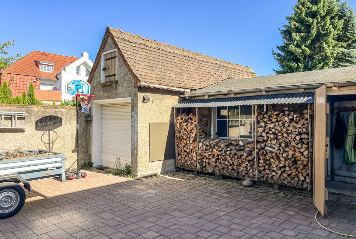 Backyard scene with neatly stacked firewood under a shed, a beige building with a tiled roof, and a basketball hoop on a wall in front.
