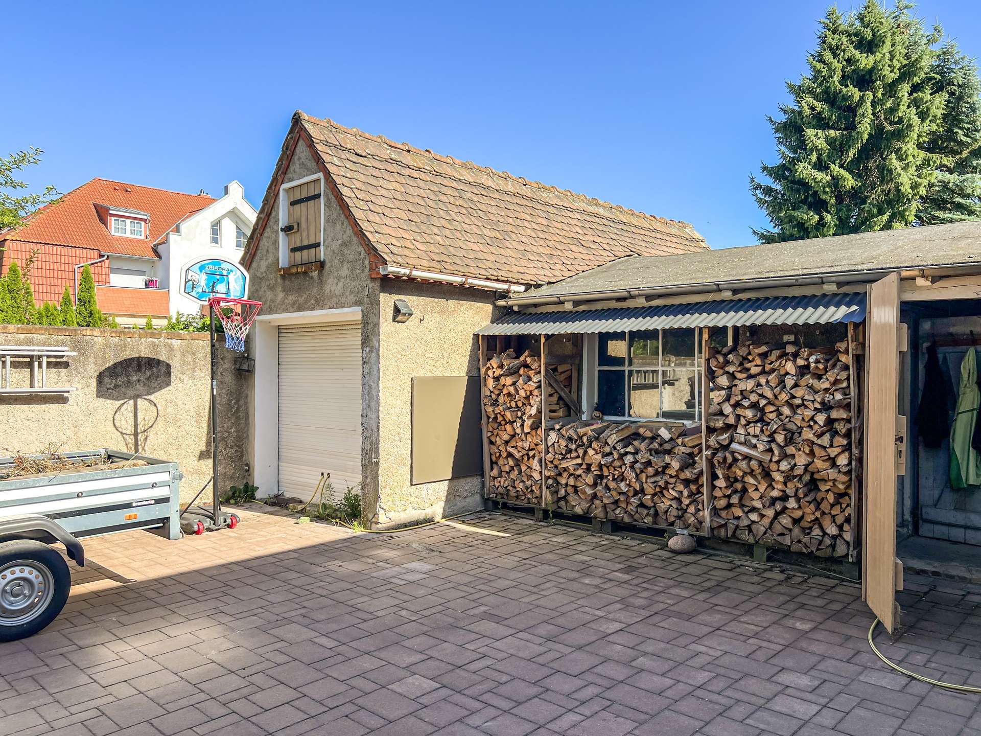 Backyard scene with neatly stacked firewood under a shed, a beige building with a tiled roof, and a basketball hoop on a wall in front.