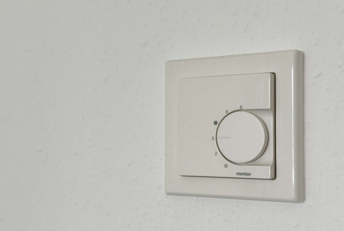 White wall-mounted thermostat with a circular dial set near 4, mounted on a pale wall