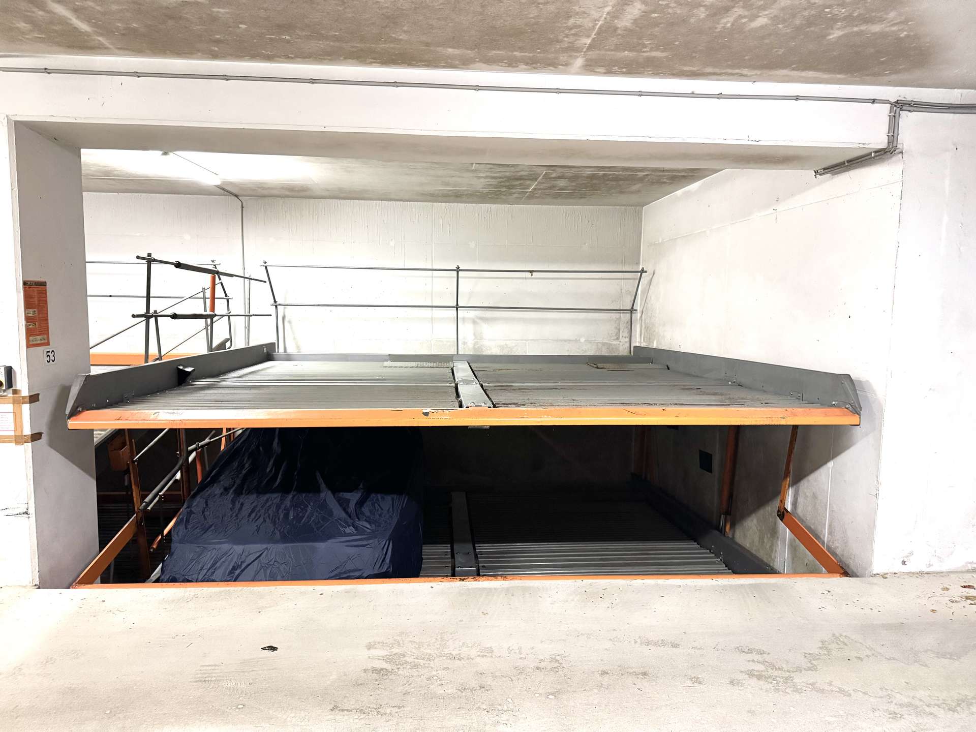 Industrial freight elevator platform with grated metal deck and orange edge rails in a white concrete loading area, scaffolding visible behind.