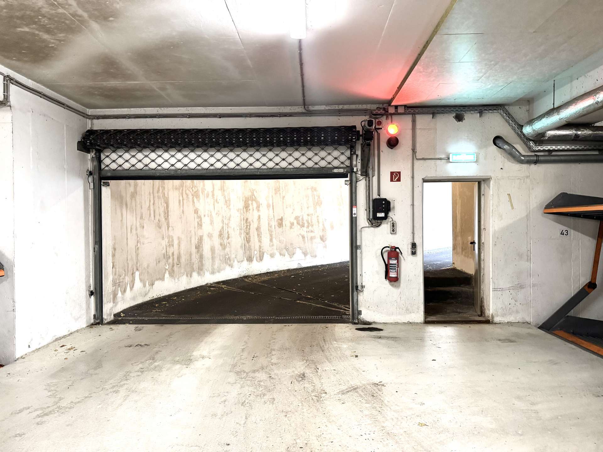 Industrial loading bay with a partially open roller door, red warning light, and a wall-mounted fire extinguisher.