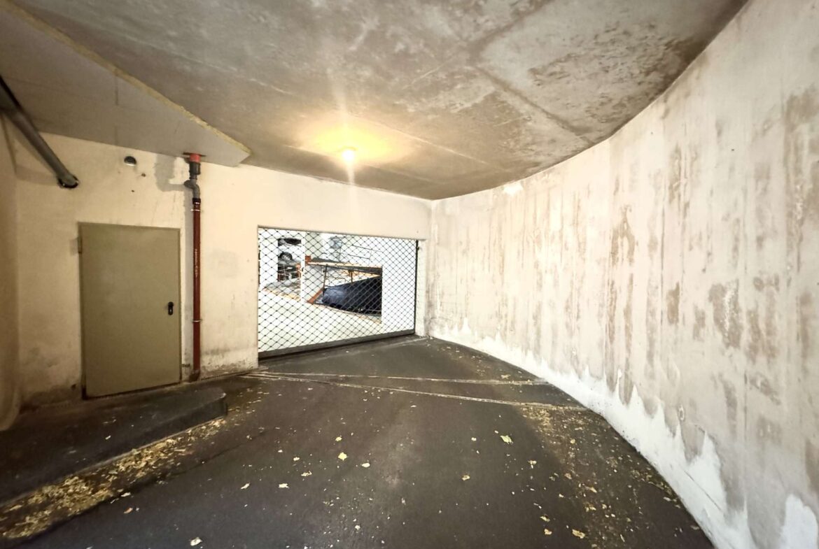Empty concrete basement with curved ceiling, white walls with stains, a red pipe along the left wall, and a small barred window at the back