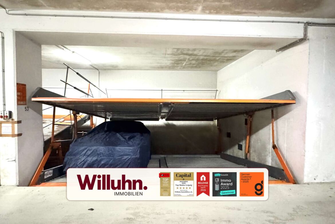 Indoor parking area with a covered vehicle under an orange-framed platform; visible Willuhn Immobilien sign and award stickers on a white panel in the foreground.