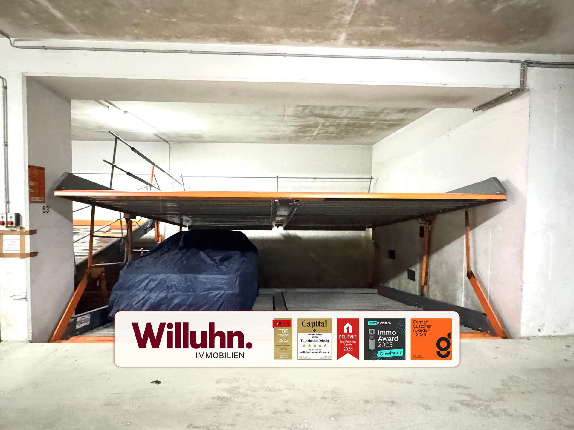 Indoor parking area with a covered vehicle under an orange-framed platform; visible Willuhn Immobilien sign and award stickers on a white panel in the foreground.