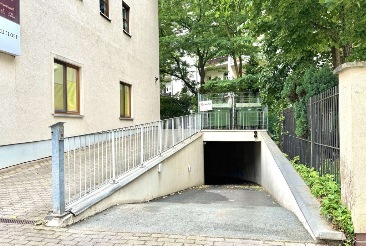 Concrete ramp with metal railing leading to a dark underground entrance beside a beige building, flanked by trees and a metal fence on the right.