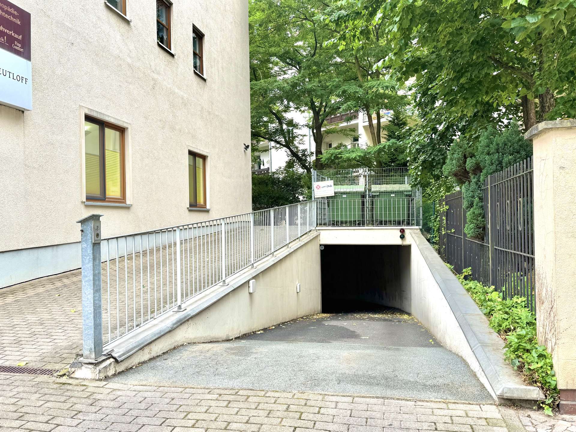 Concrete ramp with metal railing leading to a dark underground entrance beside a beige building, flanked by trees and a metal fence on the right.