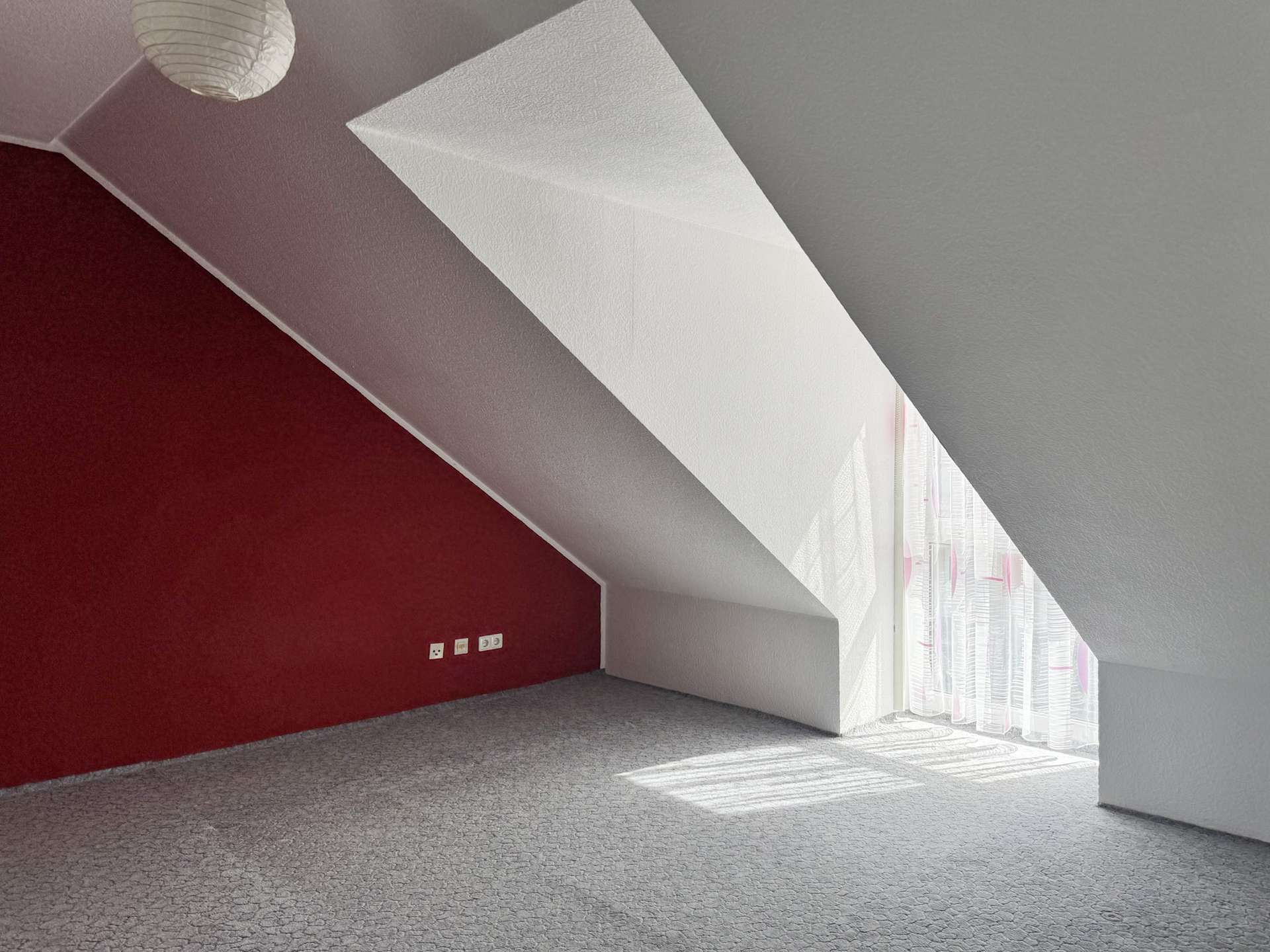 Attic-style room with a red accent wall, slanted white beam, gray walls, and sheer curtains by tall windows letting in sunlight.