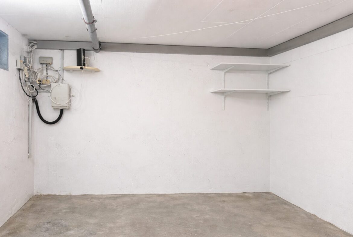 Bare basement utility room with exposed electrical equipment on the left and a white two-shelf unit on the right.