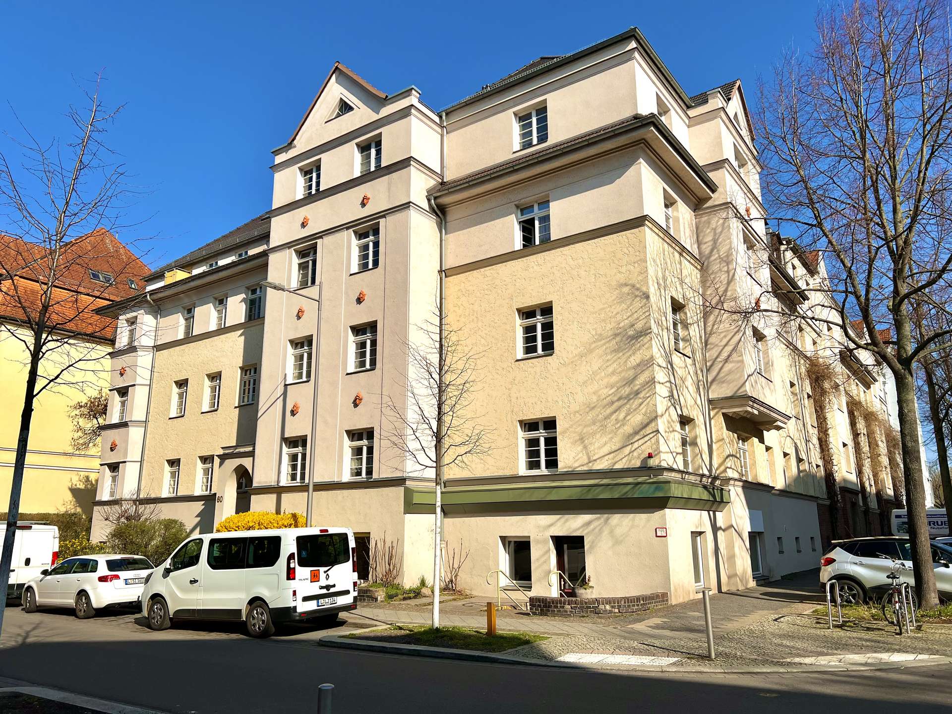 Beige multi-story apartment building with white-framed windows, leafless trees, and a clear blue sky.