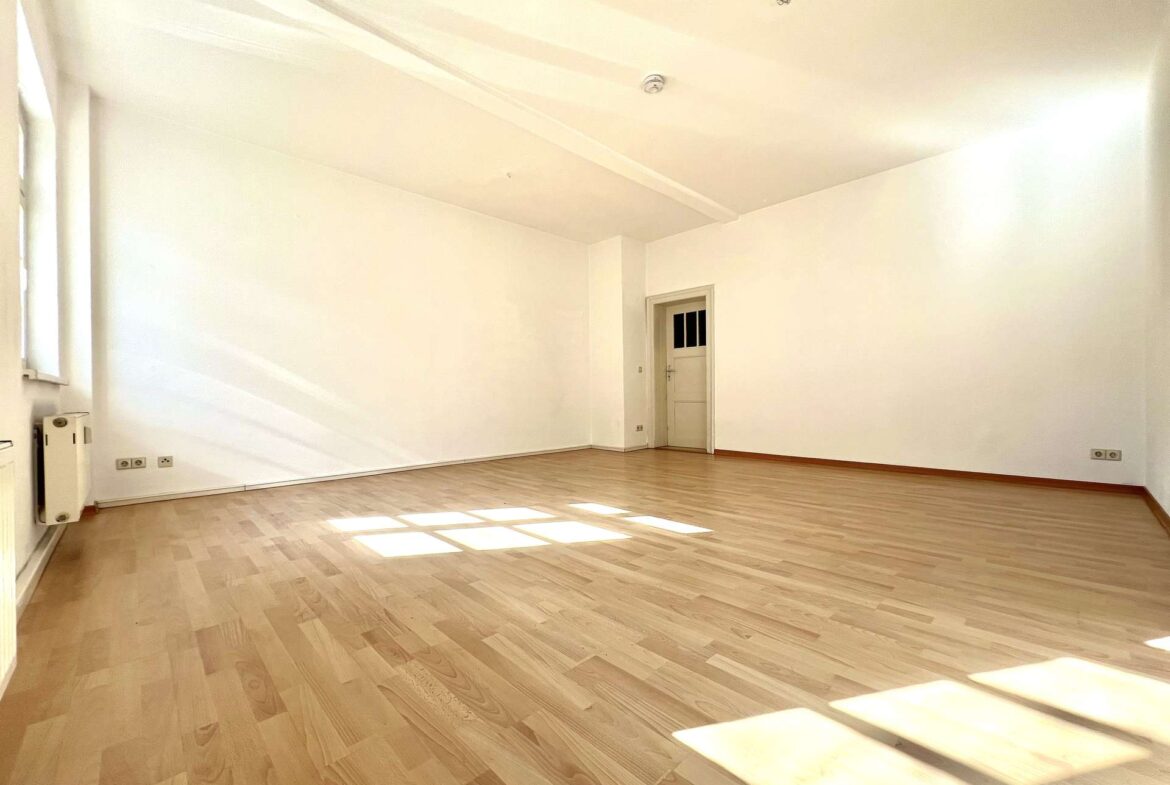 Empty sunlit room with light wood floor, white walls, a doorway at the far end, and windows casting rectangular patches of sunlight on the floor.
