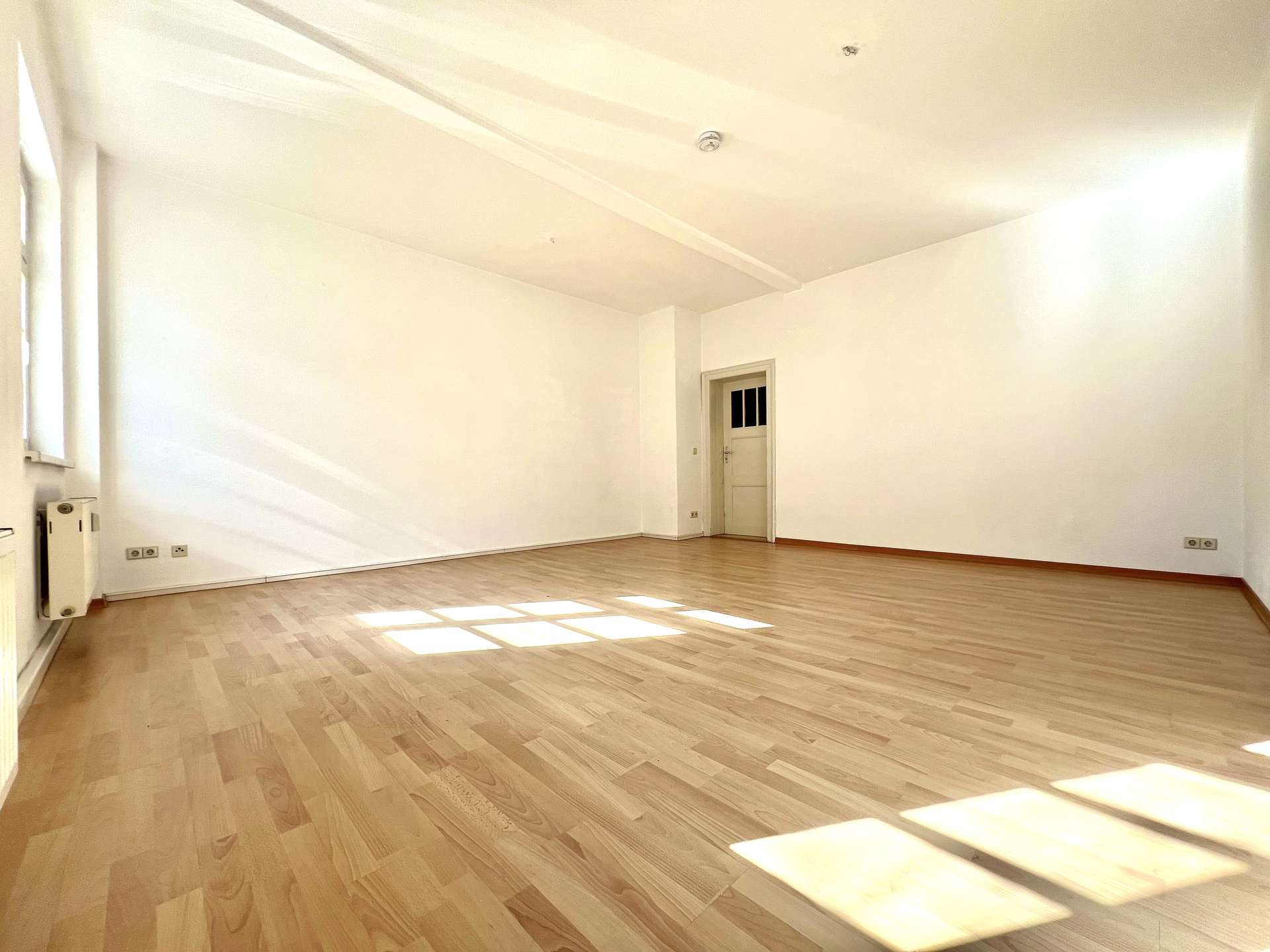 Empty sunlit room with light wood floor, white walls, a doorway at the far end, and windows casting rectangular patches of sunlight on the floor.